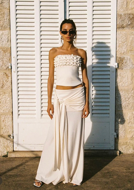 Woman in a white strapless top and flowing pants standing in front of white shutters.